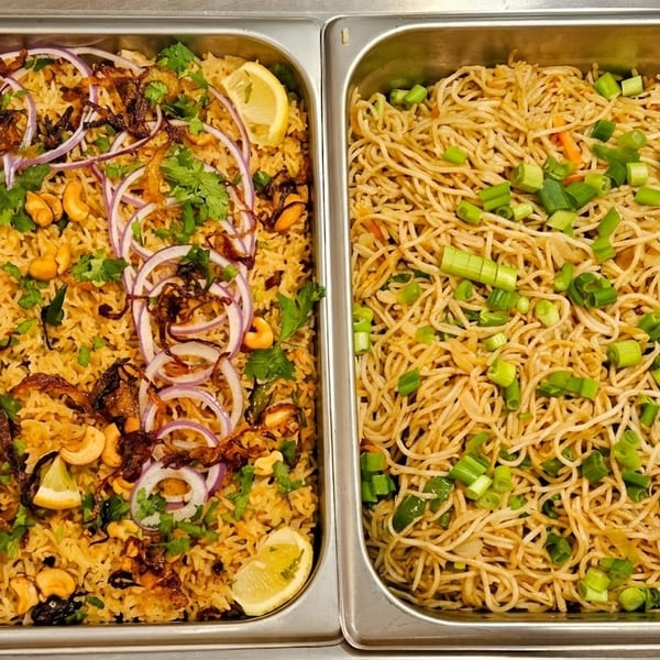 Two buffet trays, one with biryani garnished with cashews, onions, and lemon, the other with noodles and scallions.