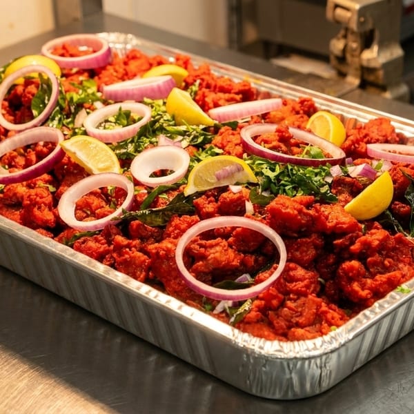 A large foil tray of bright red chicken 65, garnished with lemon wedges, red onion, and herbs.