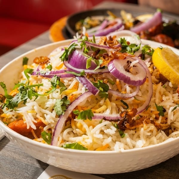 A bowl of biryani with rice, red onions, cilantro, and a lemon wedge on a wooden table.
