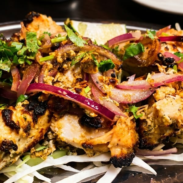 Grilled chicken tikka with red onions, cilantro, and lime on a metal platter.