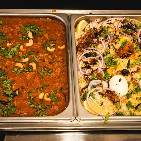 Two buffet trays, one with red curry and cashews, the other with biryani, eggs, and onions.