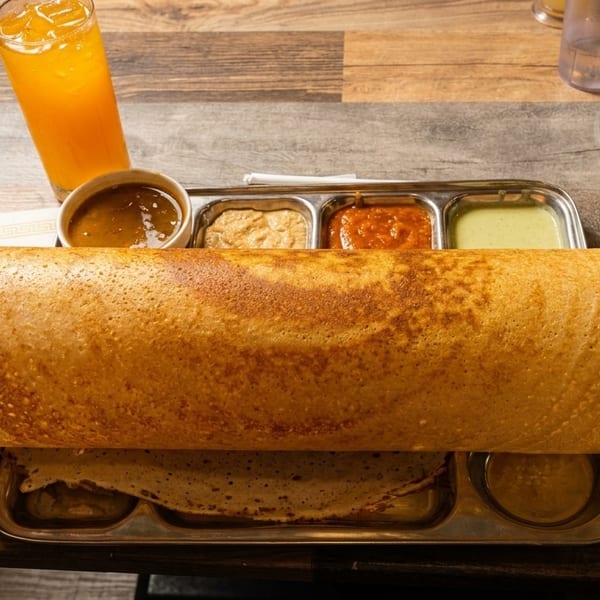 A long, golden-brown dosa served with various chutneys and a glass of orange juice on a wooden table.