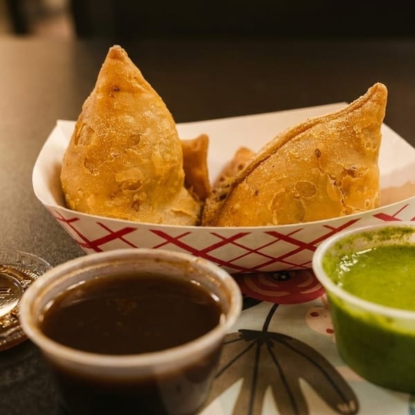 Golden-brown samosas in a paper tray, with green and brown dipping sauces on a floral placemat.